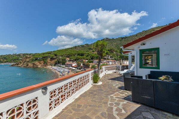 A coastal hotel with a stone-paved terrace, a white building with a green door, and a view of the beach and sea.