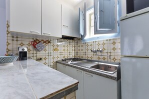 Una cocina con armarios blancos, un lavamanos de acero inoxidable, una cafetera y un ventanal con contraventanas azules.