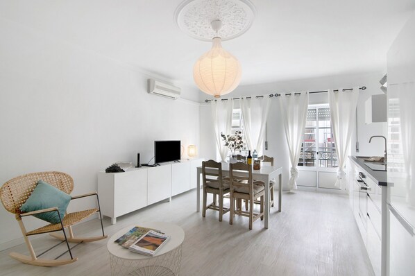 A modern living room with a dining area, white furniture, a television, and a kitchen with a window.