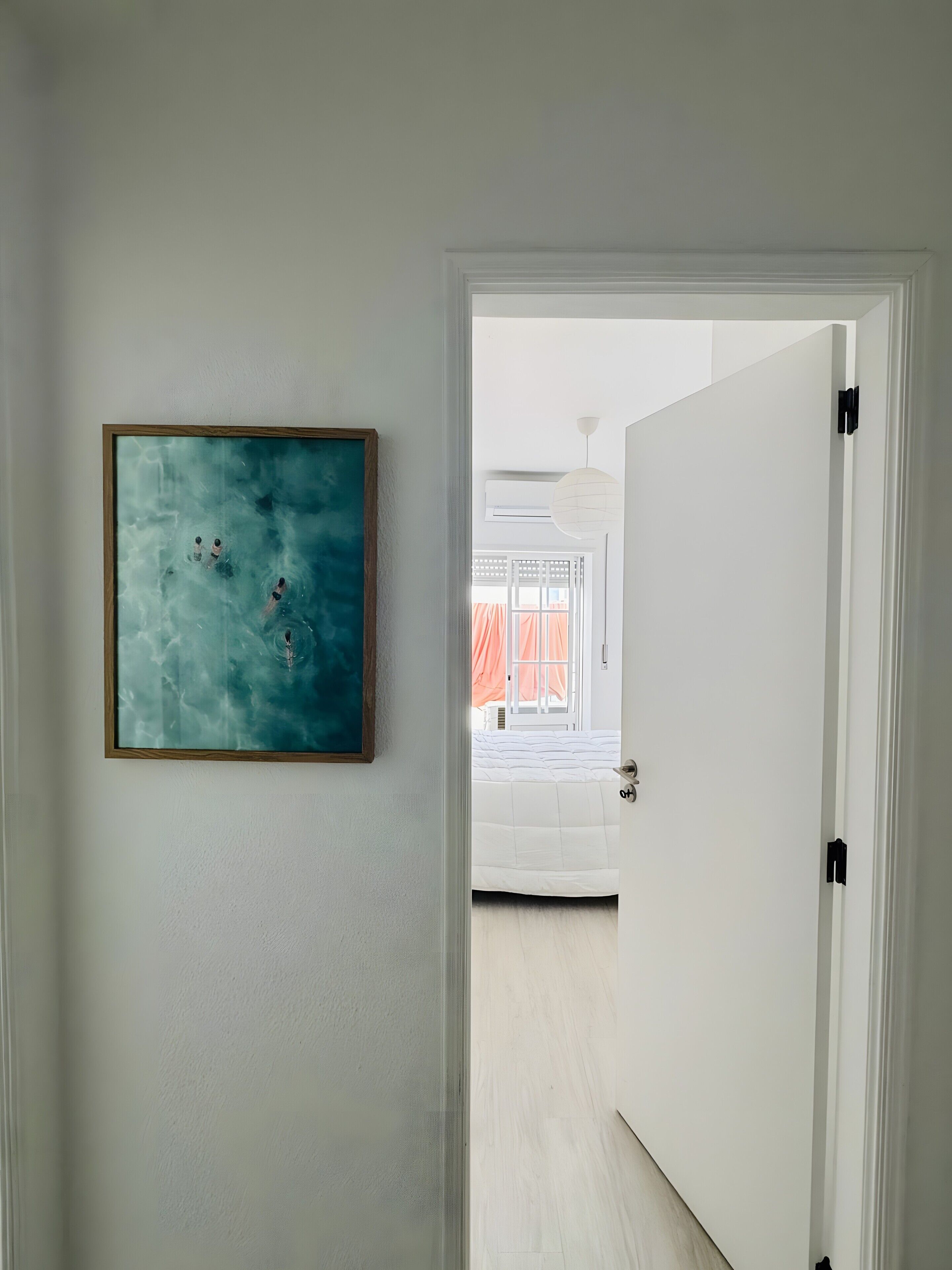 Un pasillo con un cuadro abstracto enmarcado, una puerta blanca y una vista parcial de un dormitorio con cama y cortinas.