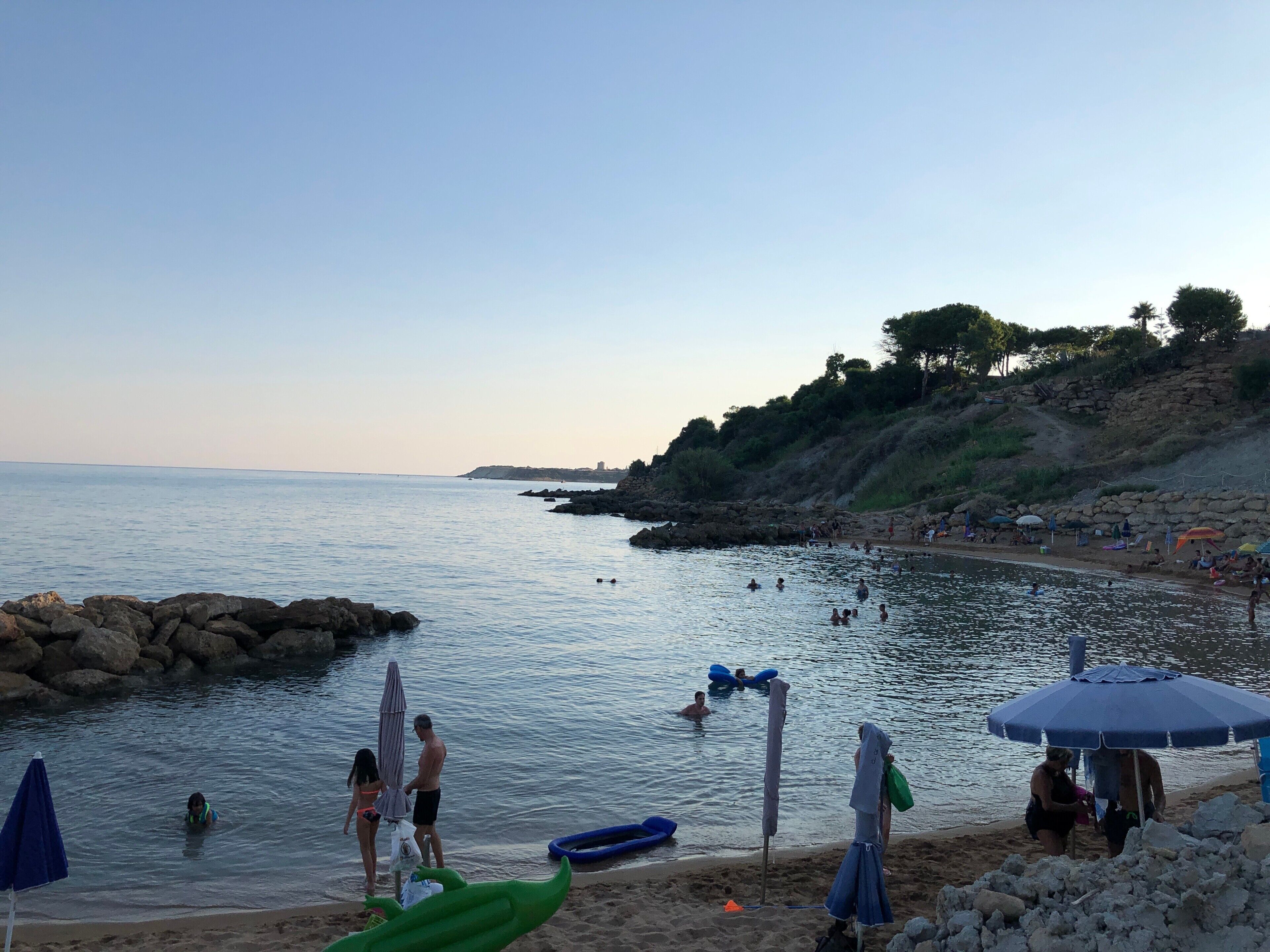 Ein Strand mit badenden Personen und Menschen am Ufer, eine felsige Küstenlinie und ein klarer Himmel.