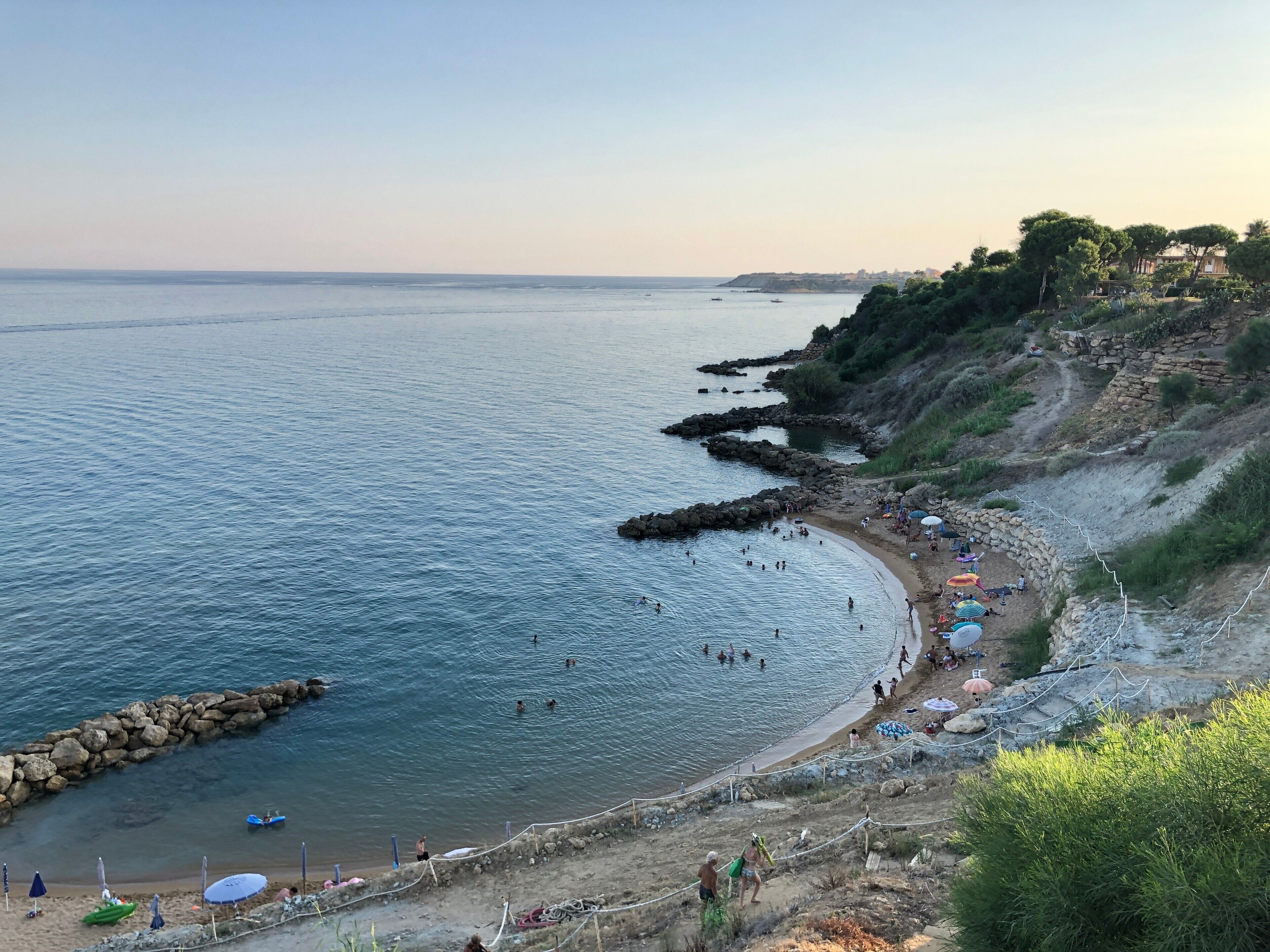 A coastal beach with a rocky shoreline, clear water, and a few umbrellas and people.