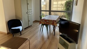 Une petite pièce bien éclairée, avec un plancher en bois, une table recouverte d’une nappe à motifs floraux, une chaise noire, un meuble et un téléviseur posé sur un support.