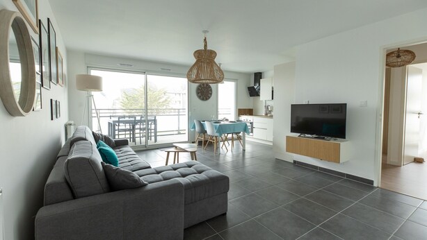 A modern living room with a grey sectional sofa, a dining area with a blue tablecloth, and a TV mounted on the wall.