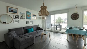 A modern living room with a grey sofa, a coffee table, and a dining area with a table set for two. There are framed pictures on the wall, a large mirror, and a ceiling light fixture.
