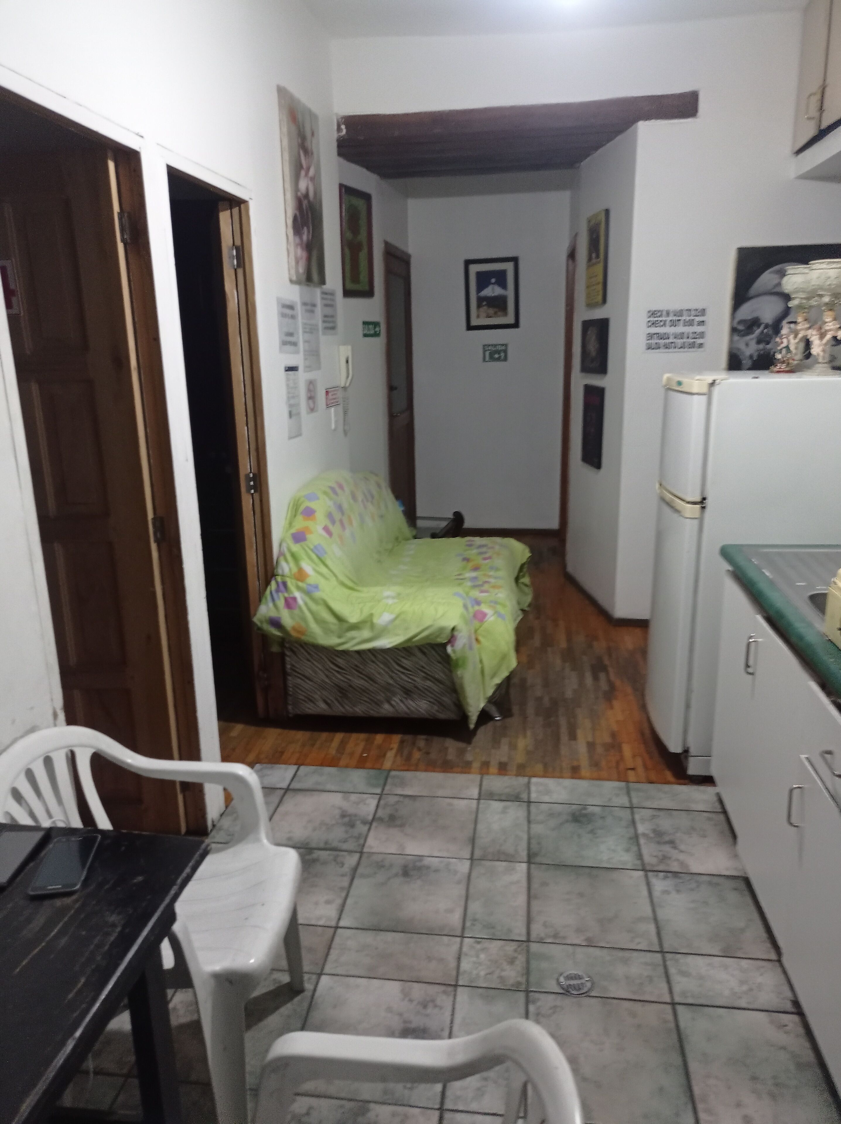 Una habitación pequeña y estrecha con una cama cubierta por un colorido edredón, una silla blanca, una mesita y un refrigerador.