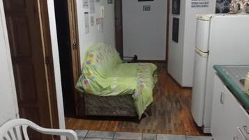 Una habitación pequeña y estrecha con una cama cubierta por un colorido edredón, una silla blanca, una mesita y un refrigerador.
