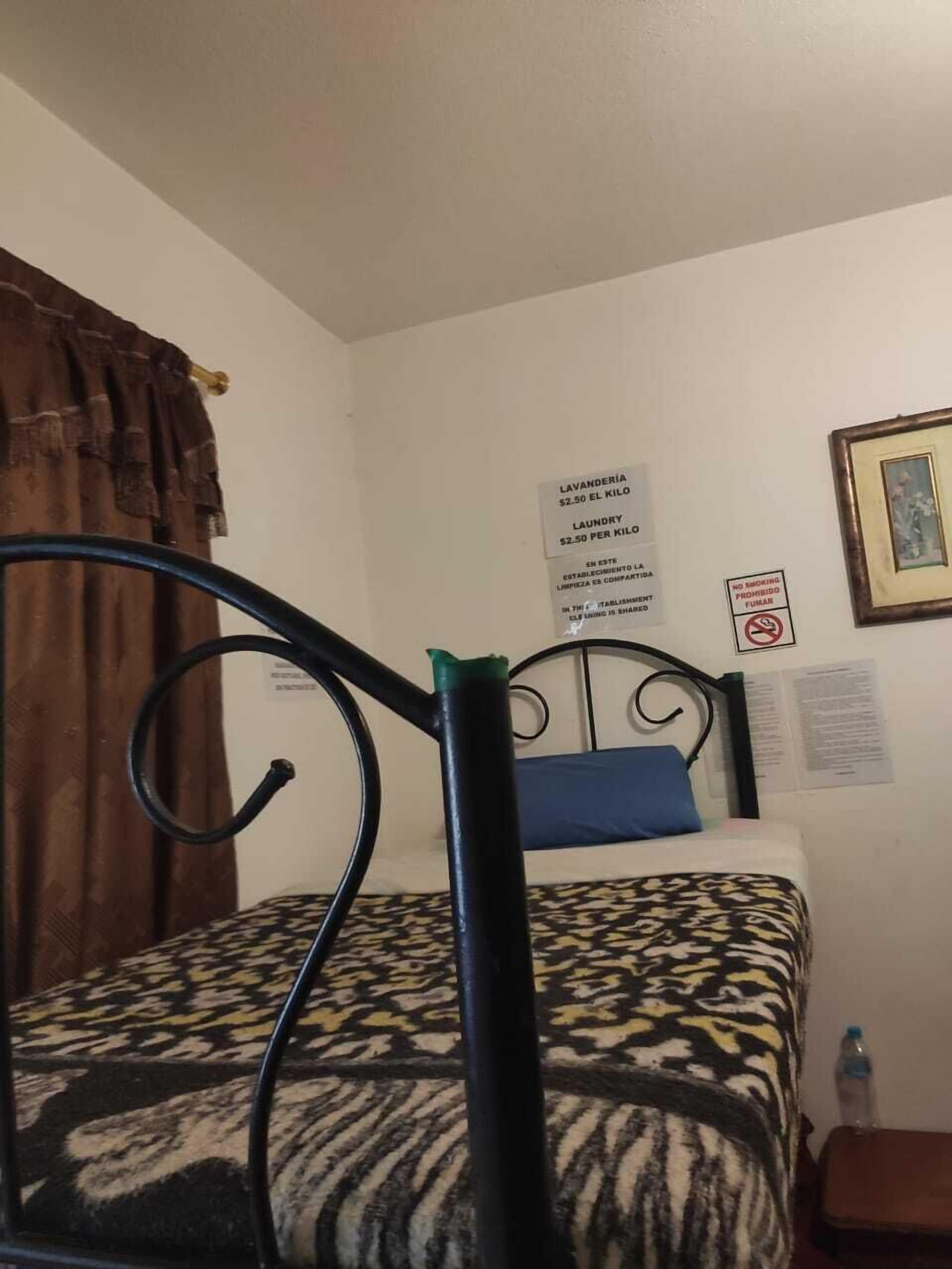 A single bed with a patterned bedspread, a metal headboard, a brown curtain, and a wall with a 'No Smoking' sign and a laundry sign.