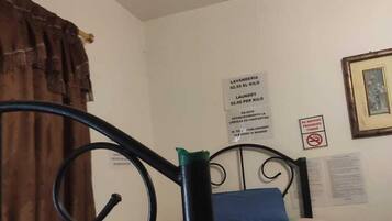 A single bed with a patterned bedspread, a metal headboard, a brown curtain, and a wall with a 'No Smoking' sign and a laundry sign.