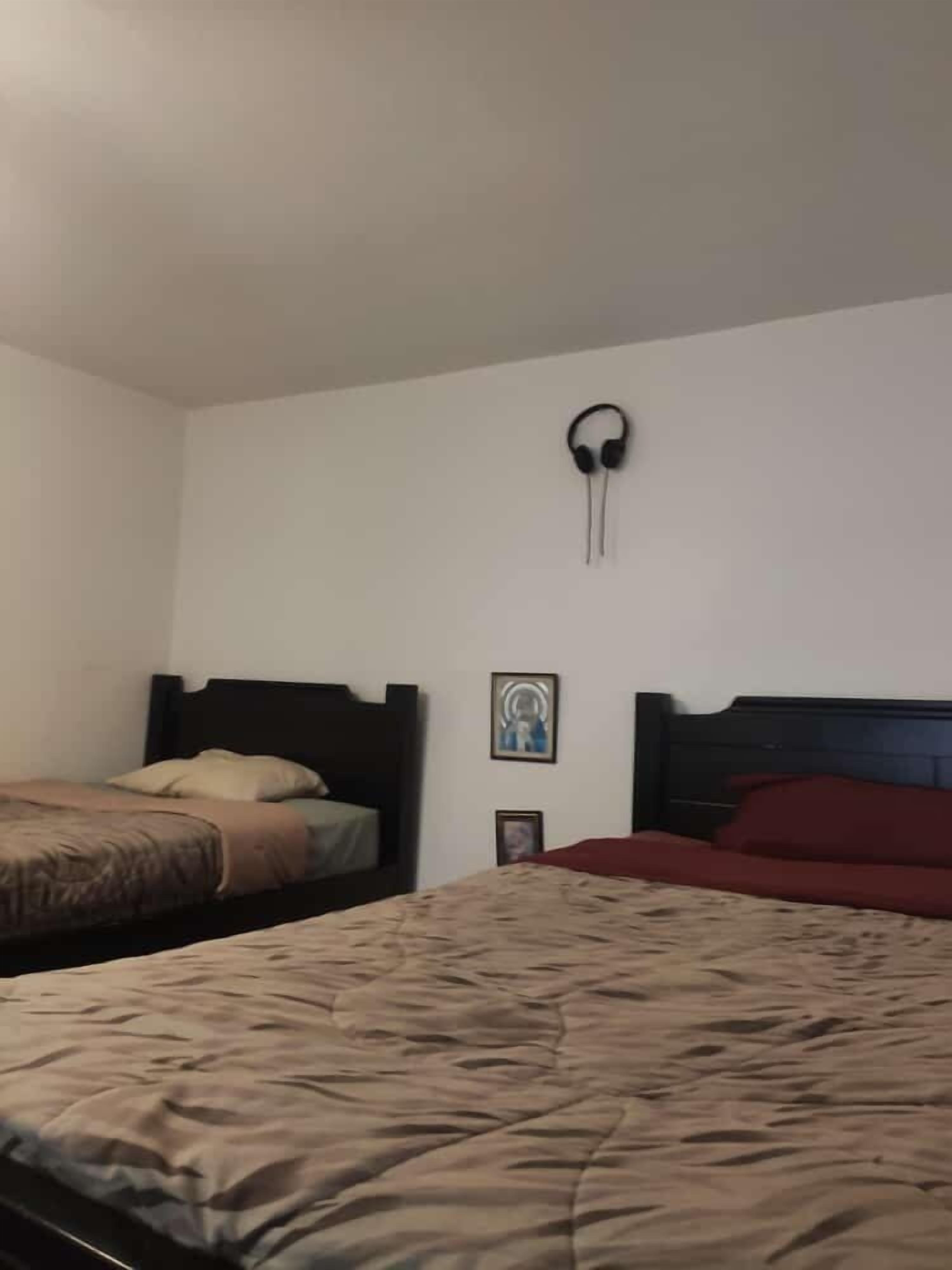 A bedroom with two beds, one with a red and one with a beige bedspread. There is a wall-mounted speaker and two framed pictures on the wall.
