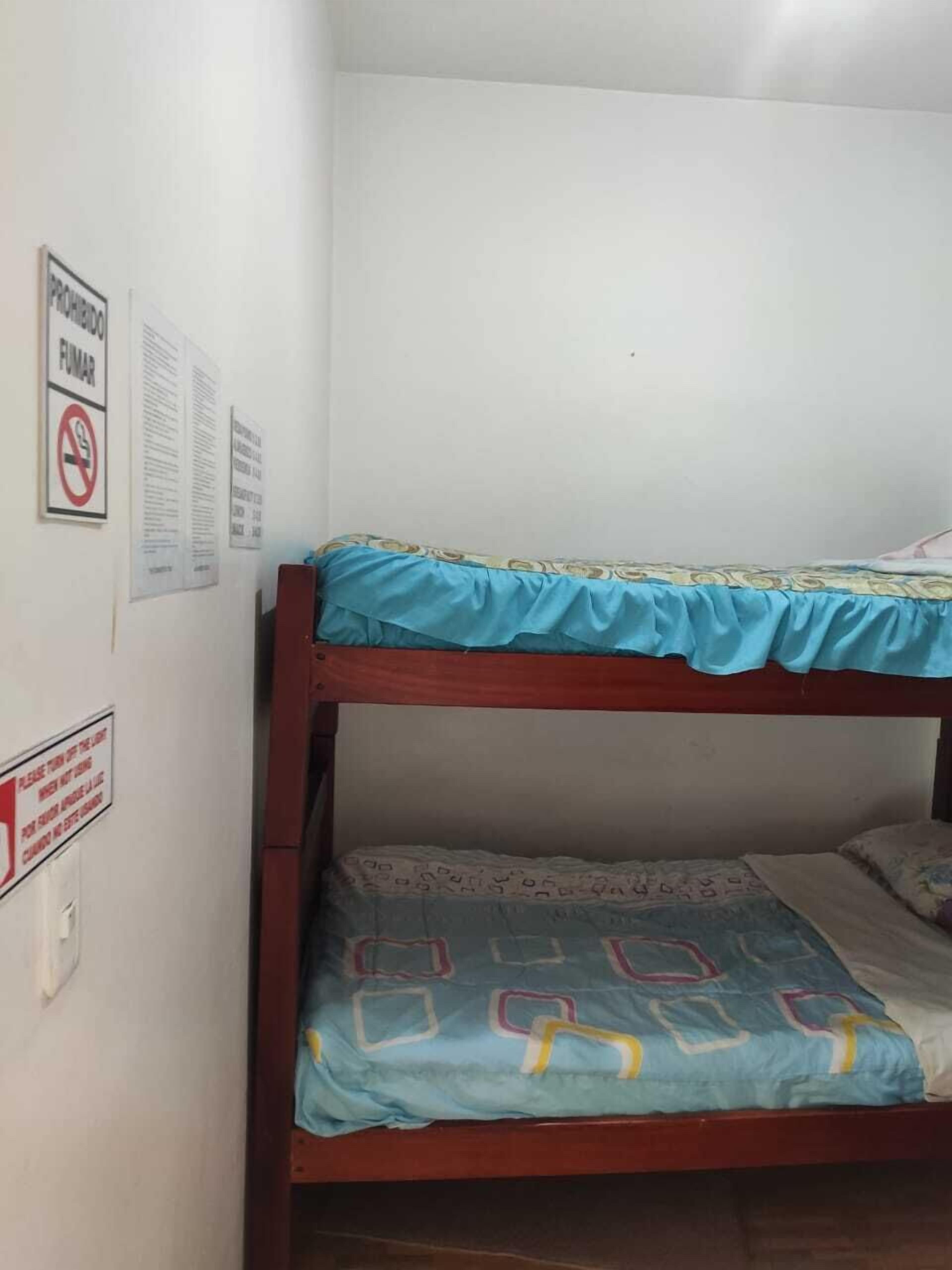 A bunk bed with a blue top bunk and a lower bunk with a patterned bedspread. There are signs on the wall, one of which has a red circle with a line through it, indicating a prohibition.