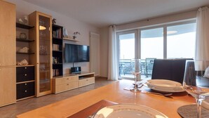Een moderne woonkamer met een houten eettafel gedekt voor twee personen, een flatscreen televisie en een balkon met uitzicht.