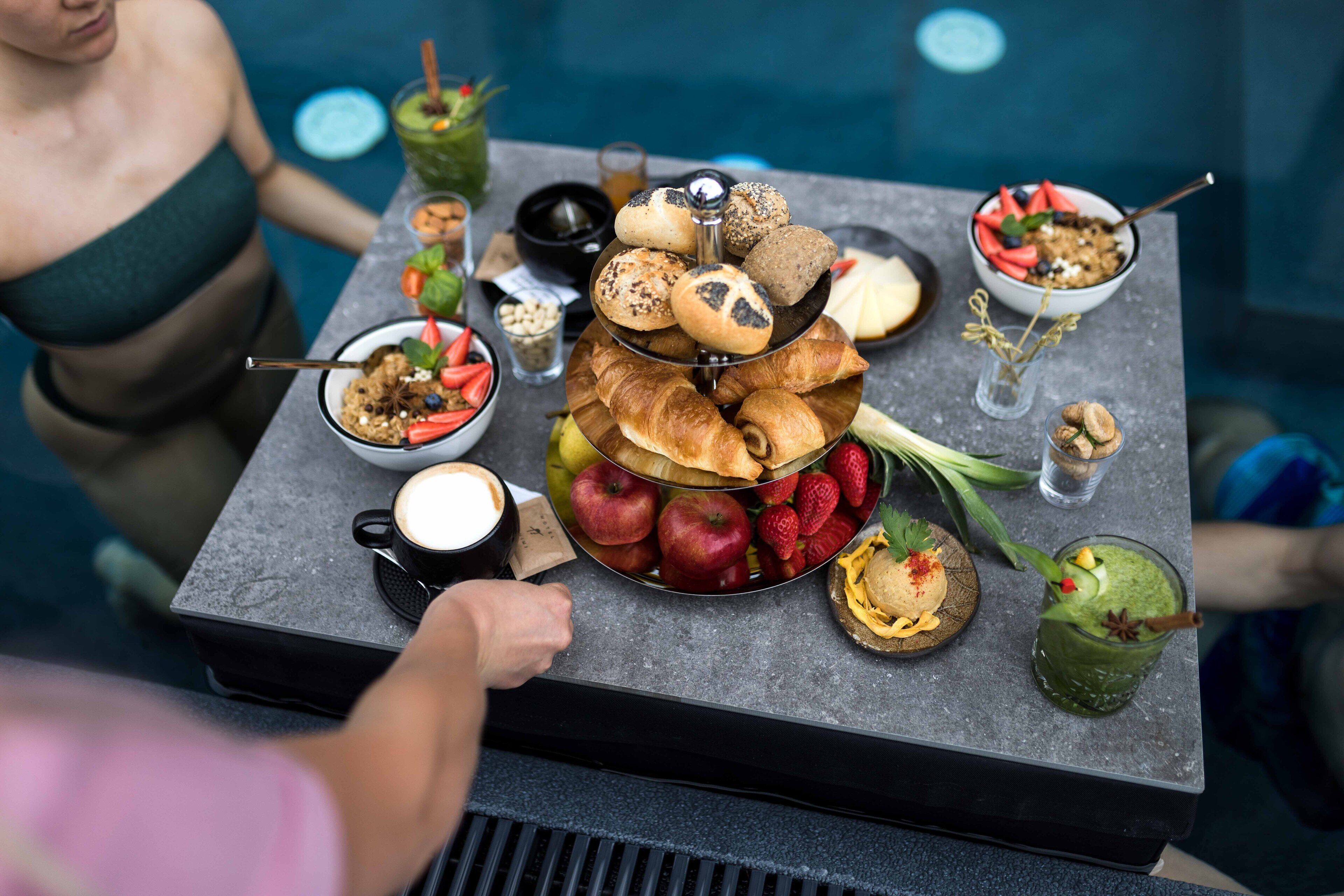 A breakfast buffet with various pastries, fruits, and beverages on a table by a pool.