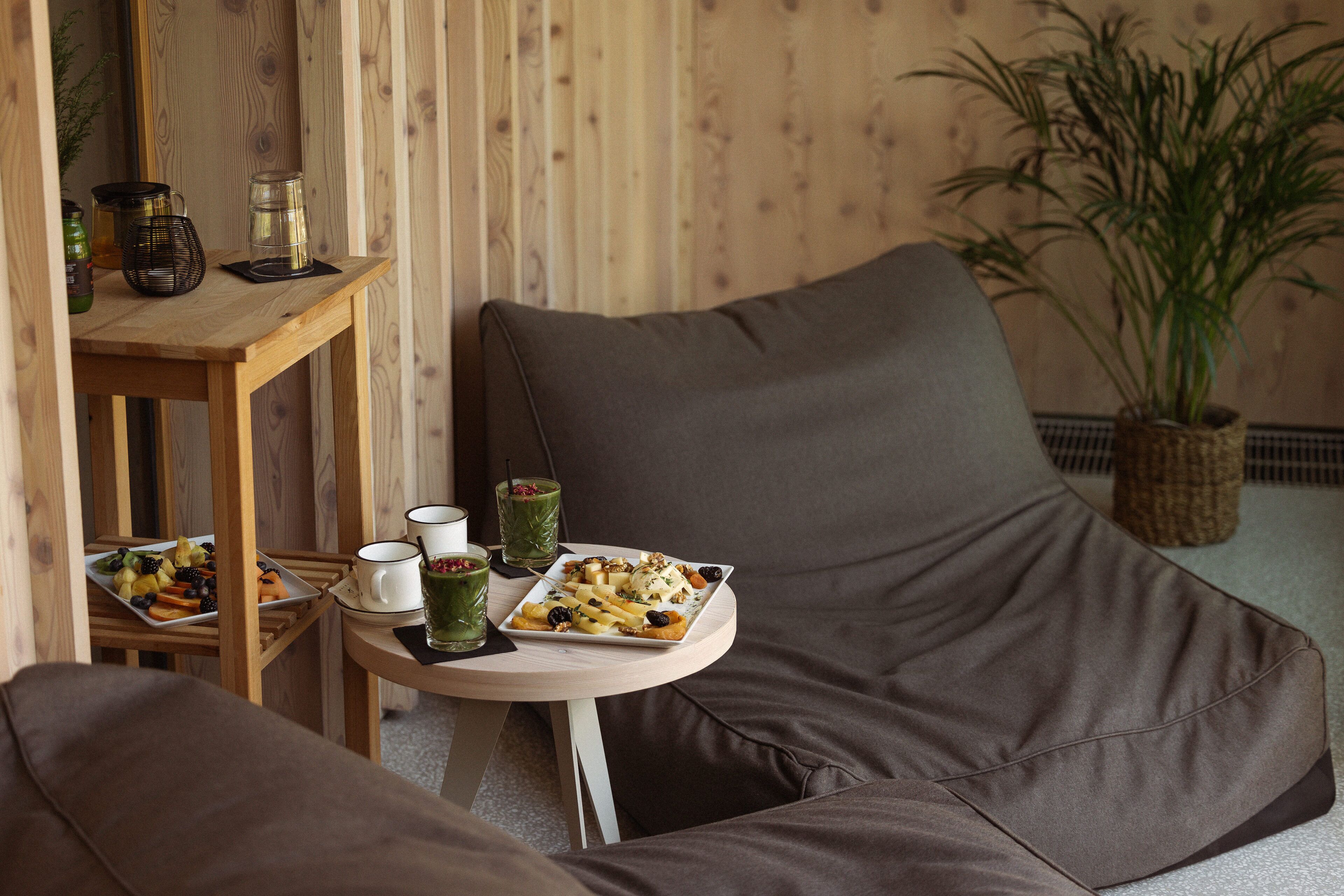 A cozy seating area with a brown sofa, a small round table with a tray of food, and a wooden side table with a green drink and a candle.