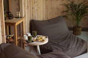 A cozy seating area with a brown sofa, a small round table with a tray of food, and a wooden side table with a green drink and a candle.