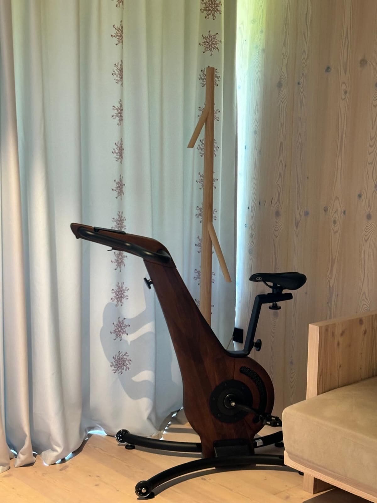 Un vélo d’appartement est installé dans une pièce dont le sol est en bois, qui contient également un canapé beige et un rideau orné d’un motif floral.