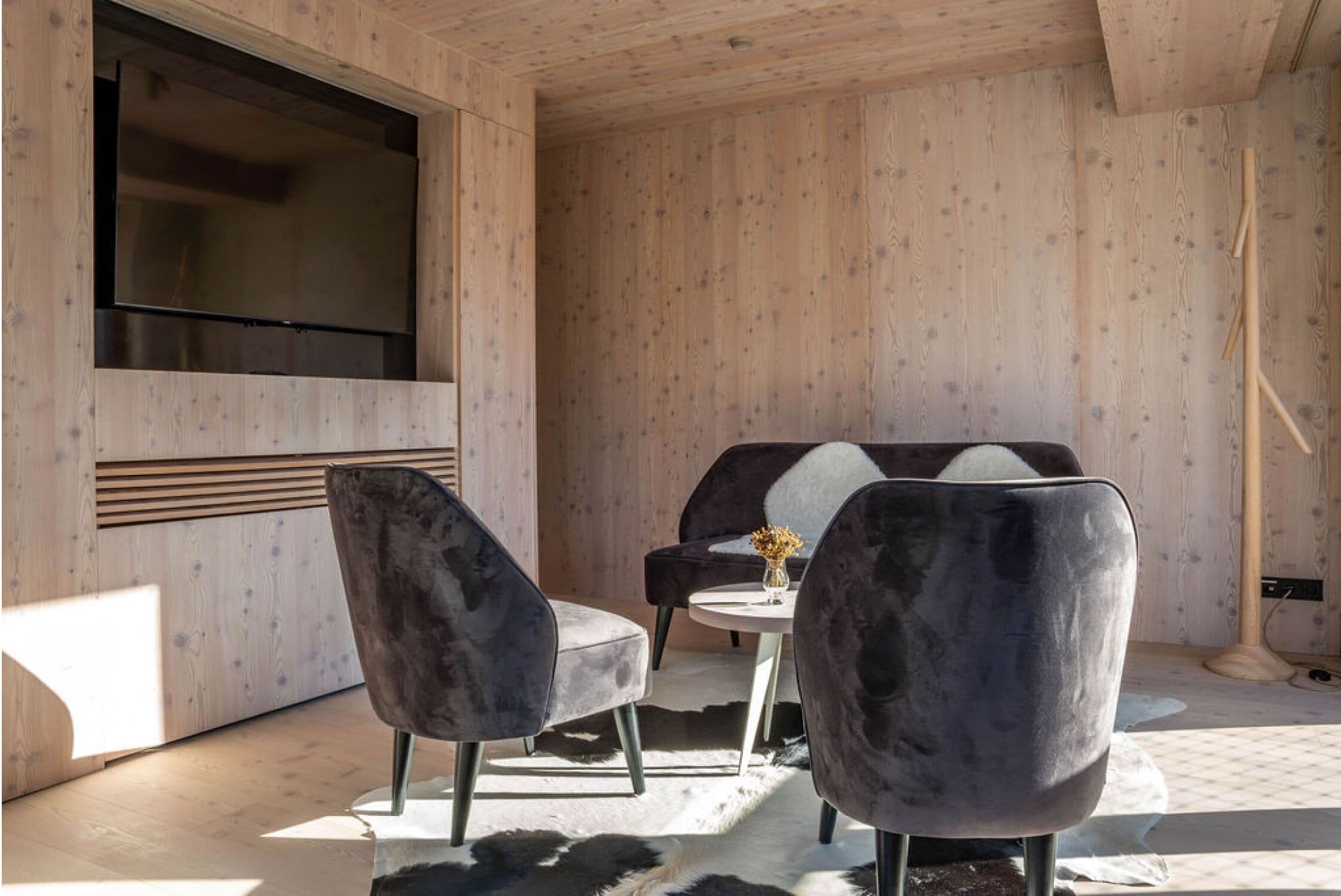 A modern room with a large flat-screen TV mounted on a wooden wall, two grey upholstered chairs, a small round table with a vase, and a large window allowing natural light.