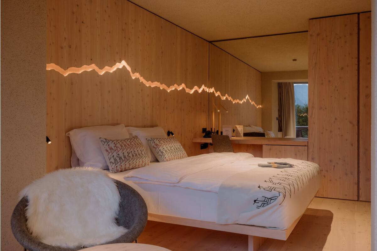 A modern bedroom with a wooden wall, a bed with patterned bedding, a fluffy chair, and a bedside table with a lamp.