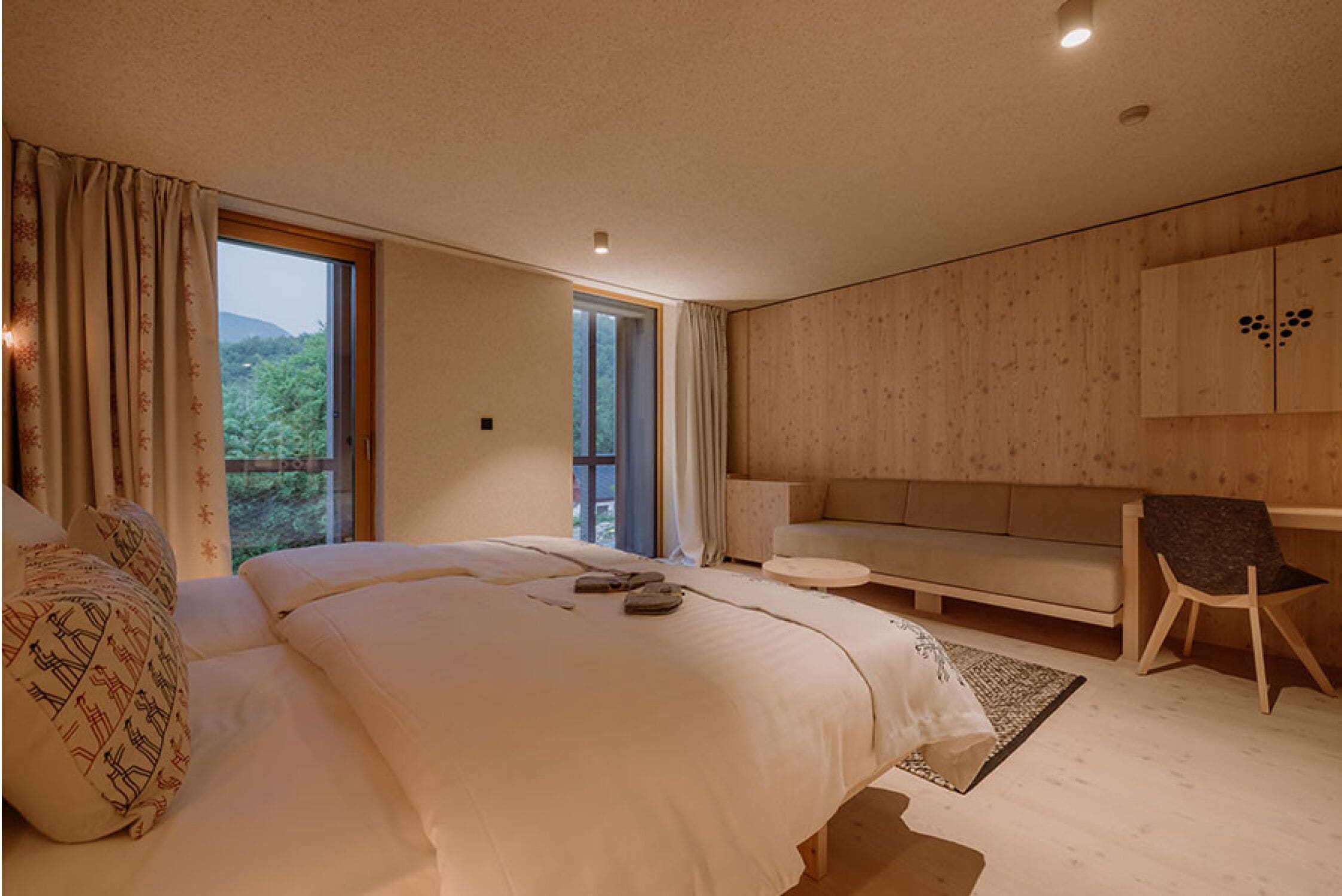 A modern bedroom with a large bed, a sofa, and a chair. There is a window with curtains, a wooden wall, and a ceiling light.