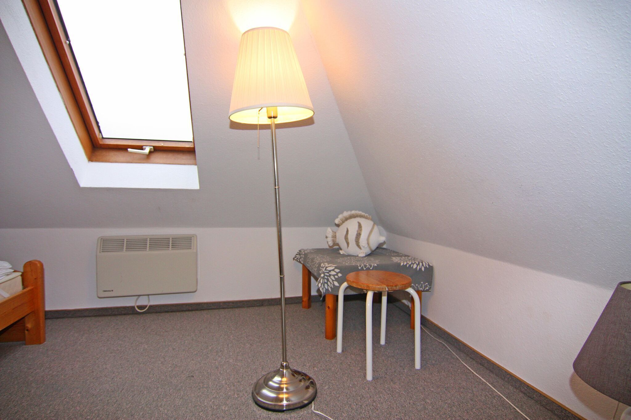 Una habitación pequeña y bien iluminada con tragaluces, una lámpara de pie, un calentador, una mesita con un cojín y una cama.