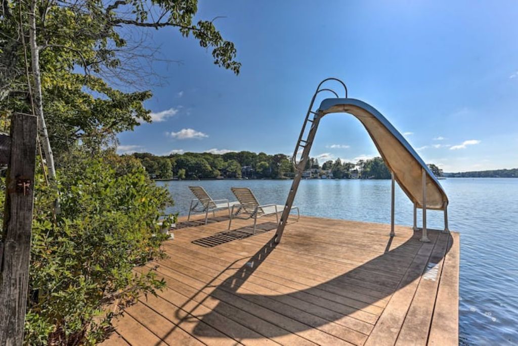 A wooden deck with a metal swing chair and two lounge chairs facing a calm lake.