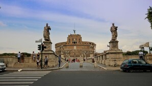 A view of the Colosseum with two large statues on either side, a pedestrian crossing, and a road leading to the entrance. - Two Bedroom Apartment (ROMA)