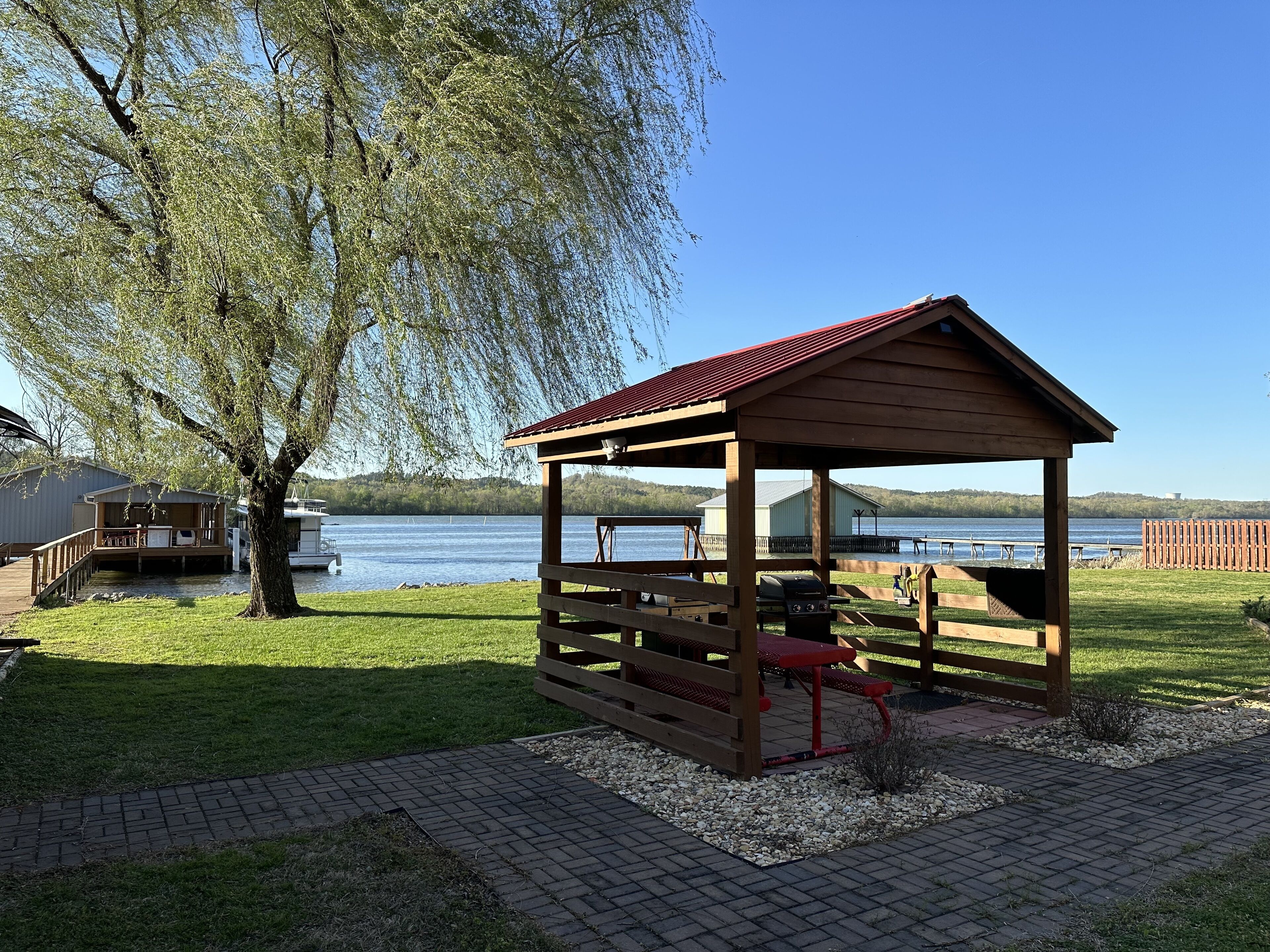 A wooden gazebo with a red roof overlooking a lake, surrounded by a grassy area and a tree.