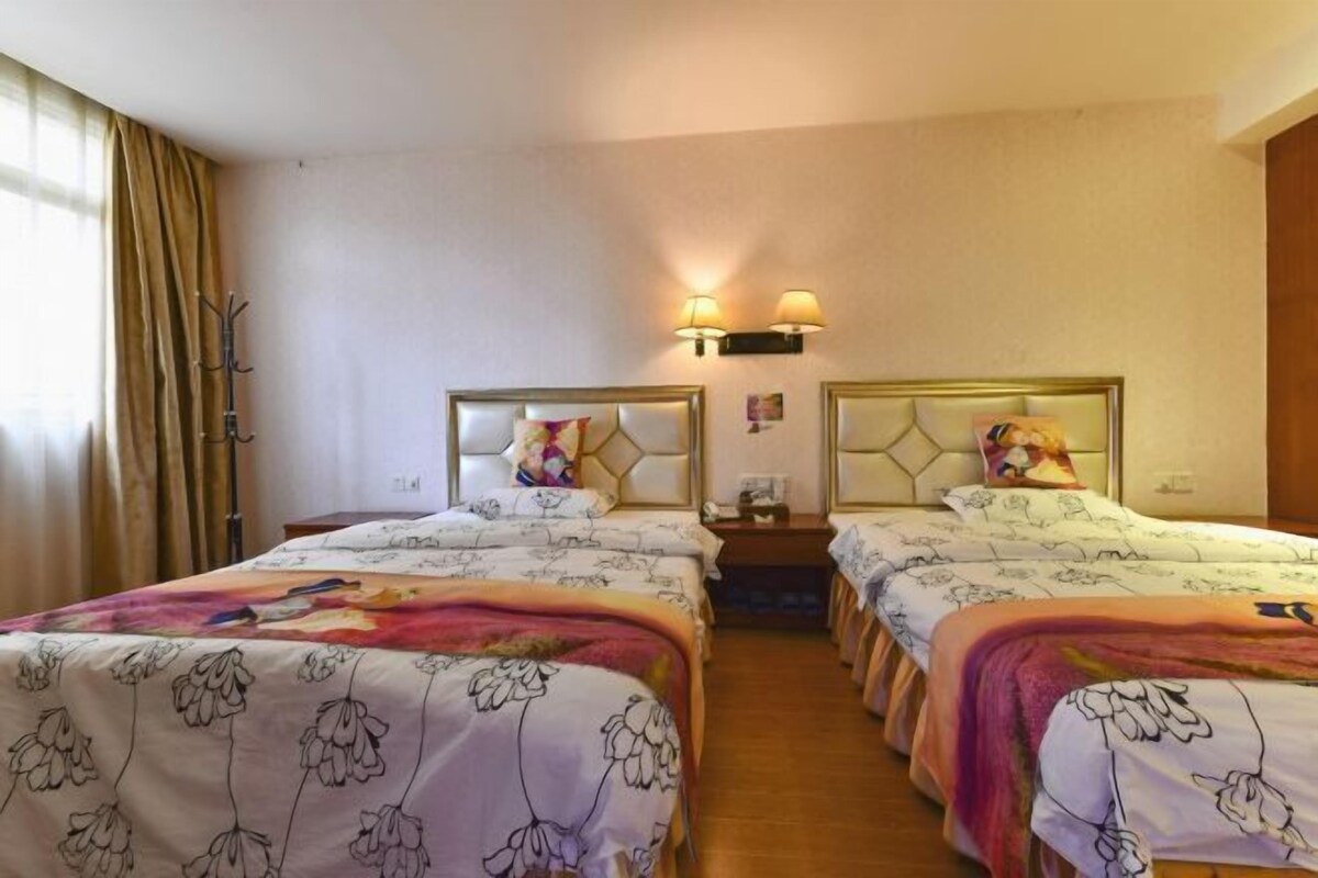 A hotel room with two beds, each with floral-patterned bedding and headboards. There are wall-mounted lamps and a coat rack.