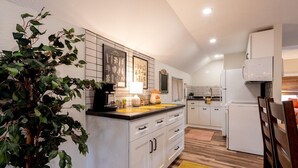 A modern kitchen with white cabinets, a central island, and a dining area with wooden chairs. - The Back Yard-Upper Near Downtown LG |Balcony |Yard Now Dog Friendly (Lake Geneva)