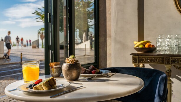 A breakfast table with a glass of orange juice, a plate with a slice of cake and strawberries, a small vase with dried flowers, and a bowl of fruit. - Pousada Alfama (Lisbon)