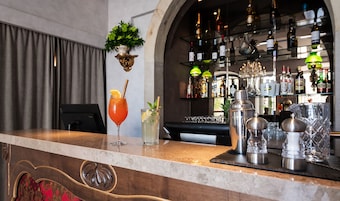 Uma área de bar com balcão, duas bebidas, um coqueteleira e uma adega ao fundo.