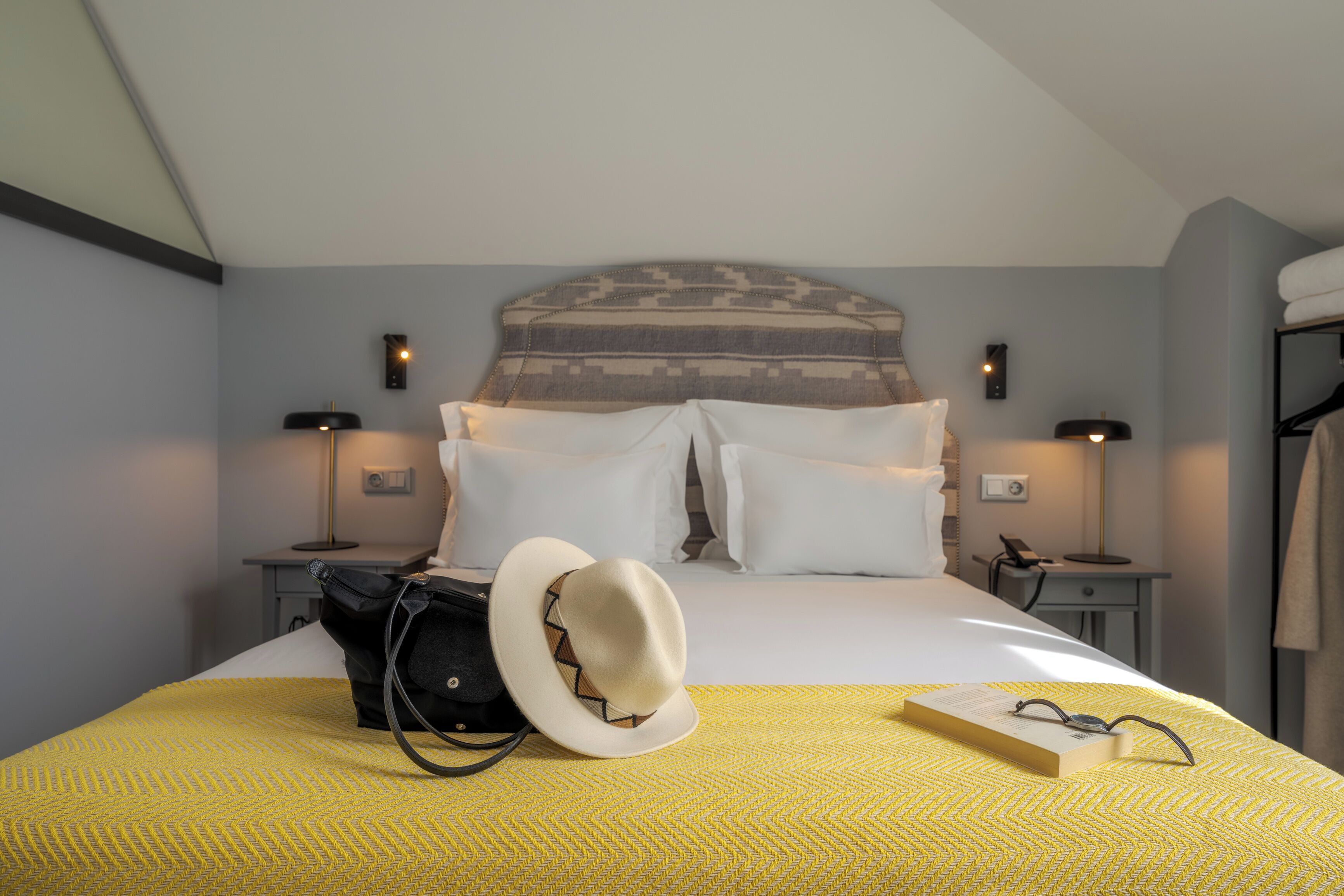 A neatly made bed with white linens, a yellow patterned bedspread, and a white hat on the bed. There are two bedside tables with lamps, a black bag, and a book with glasses on the bed.