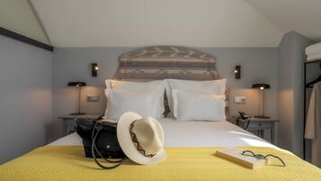 A neatly made bed with white linens, a yellow patterned bedspread, and a white hat on the bed. There are two bedside tables with lamps, a black bag, and a book with glasses on the bed.