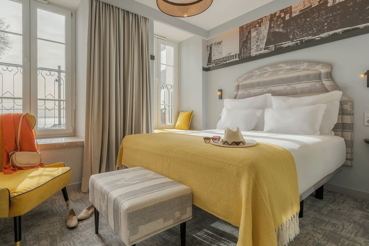 Une chambre d’hôtel comprenant un lit, une chaise, un banc, une fenêtre avec des rideaux, une œuvre d’art accrochée au mur et une lampe.