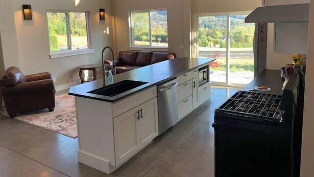 A modern kitchen with a central island, white cabinetry, and a black stove. There are large windows providing natural light and views of the outdoors. - Luxury shome, views above Skagit Valley- EV charge (Mount Vernon)
