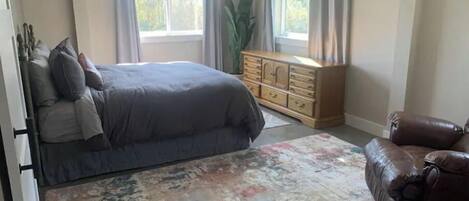A bedroom with a bed, dresser, chair, and large windows.