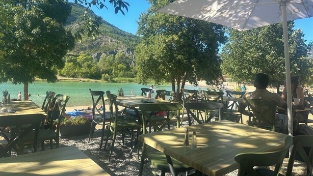 Un área de comedor al aire libre con mesas y sillas, rodeada de árboles y con vista a un cuerpo de agua.