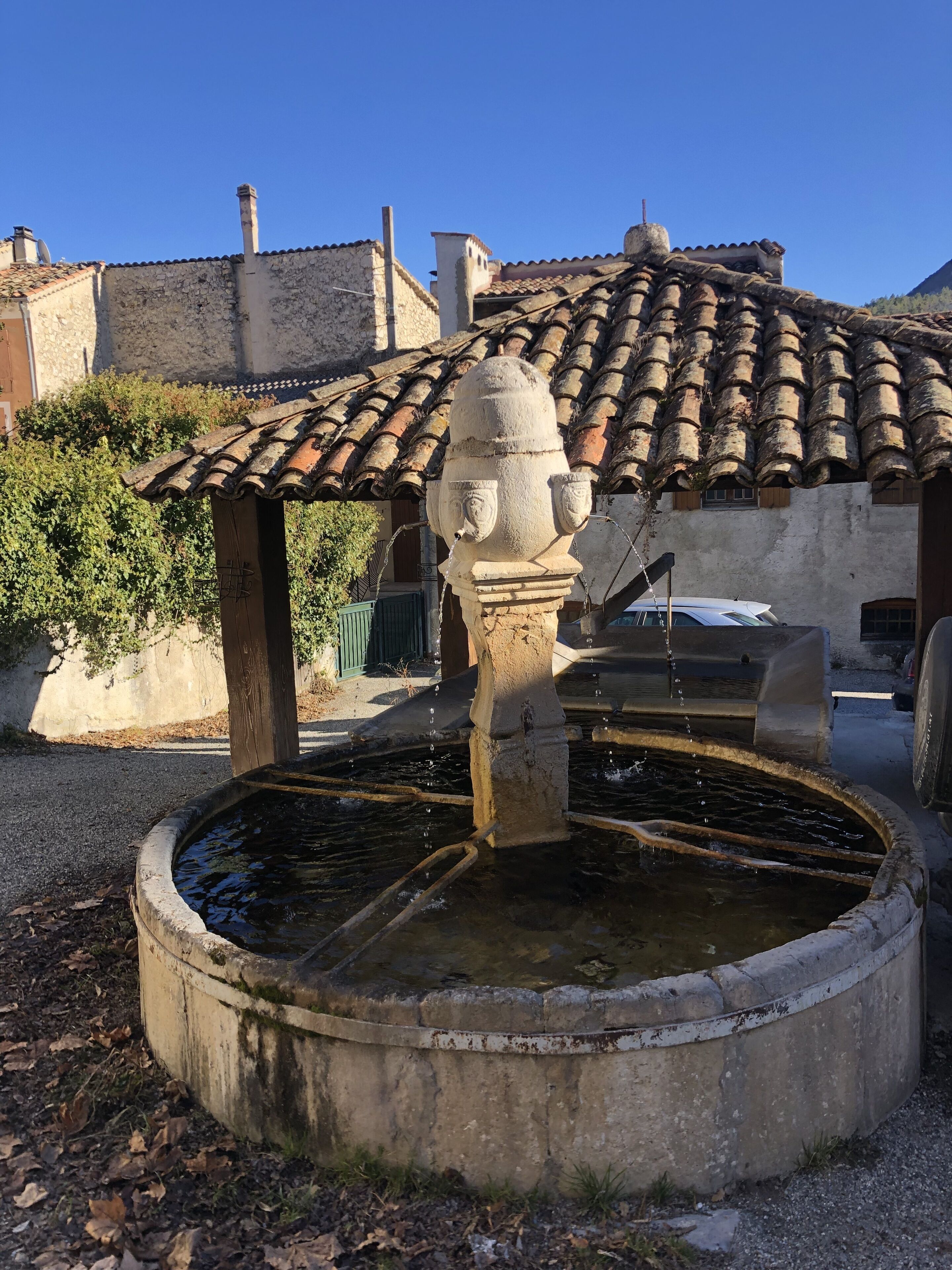 Une fontaine traditionnelle en pierre, dotée d’un bassin circulaire, entourée d’un mur de pierre et surmontée d’une structure de toit en tuiles.