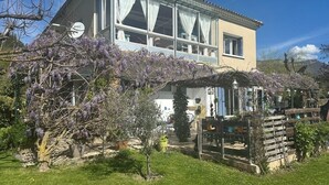 Una casa de dos pisos con un balcón, rodeada por un jardín con árboles de flores moradas.