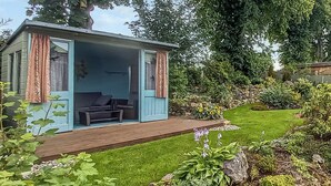 A cozy cabin with a wooden deck, a seating area with cushions, and a garden with various plants and flowers. - 3 bedroom accommodation in Biggar (Biggar)