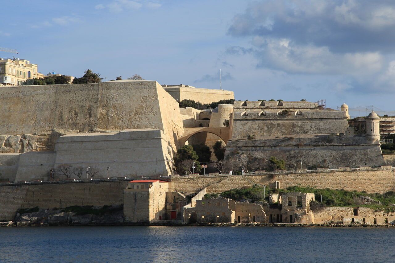 Una estructura histórica fortificada con un gran arco de entrada, muros de piedra y una masa de agua al frente.