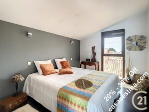 A bedroom with a bed, pillows, a nightstand, a lamp, a decorative mirror, and a window with a view of buildings.