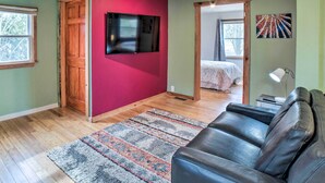 Una habitación con una puerta de madera, un televisor de pantalla plana montado en una pared roja, un sofá de cuero negro y una alfombra estampada sobre el suelo de madera.