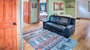 Una sala de estar con puerta de madera, sofá de cuero negro, alfombra estampada y una cama al fondo.