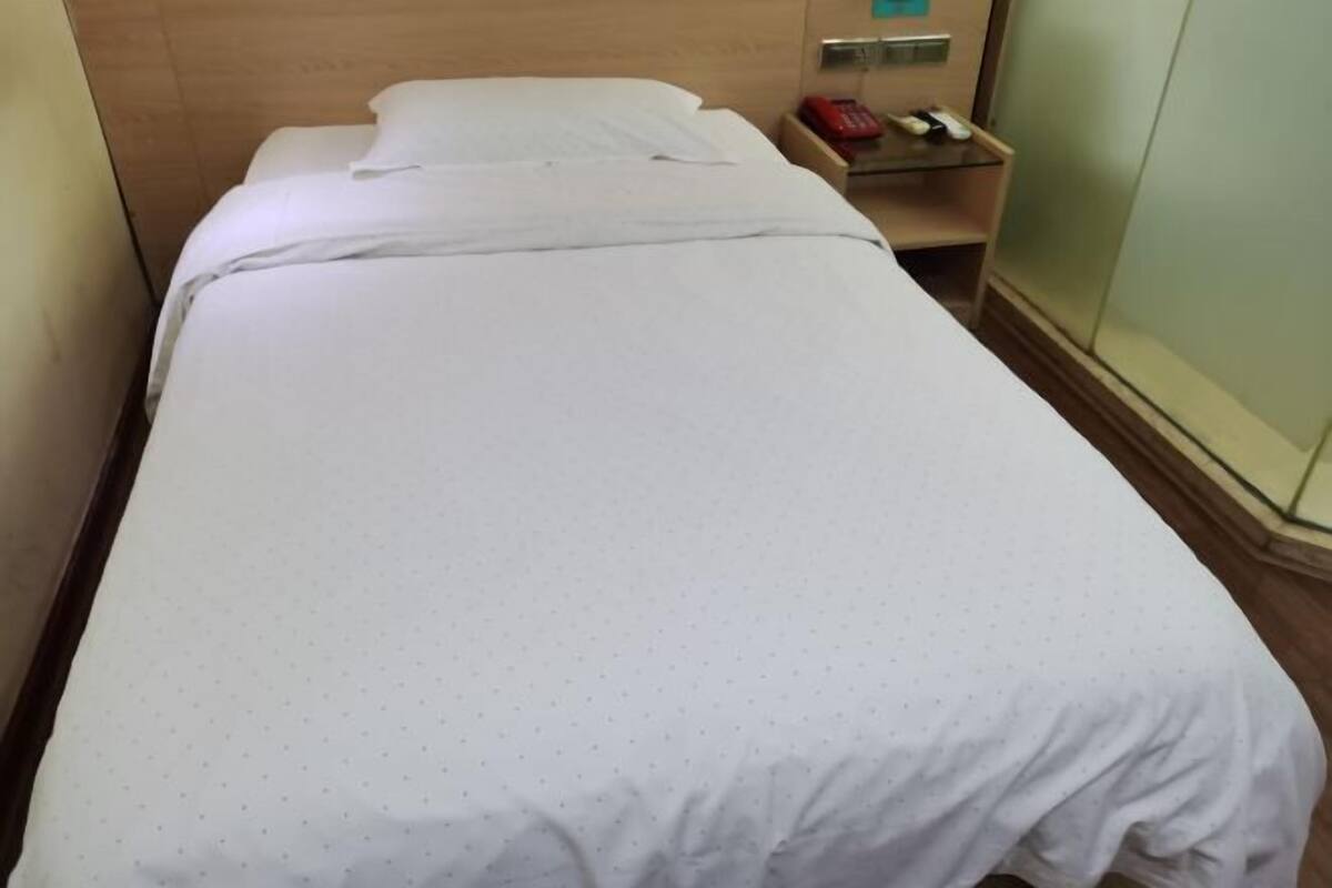 Un lit simple avec une couvre-lit blanche, une tête de lit en bois, une table de chevet avec un téléphone et un interrupteur fixé au mur.
