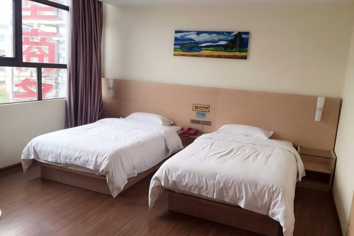 Une chambre d’hôtel avec deux lits, une fenêtre avec des rideaux, un tableau au mur et un plancher en bois.