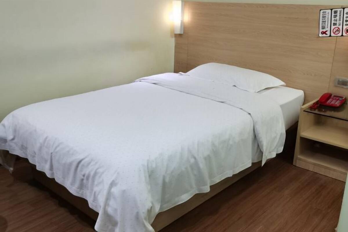 Un lit simple avec une couvre-lit blanche, une tête de lit en bois, une table de chevet avec un téléphone rouge et une lampe fixée au mur.