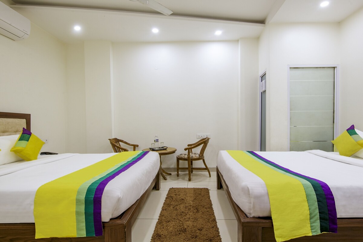 A hotel room with two beds, a small table with chairs, a door, and an air conditioning unit.