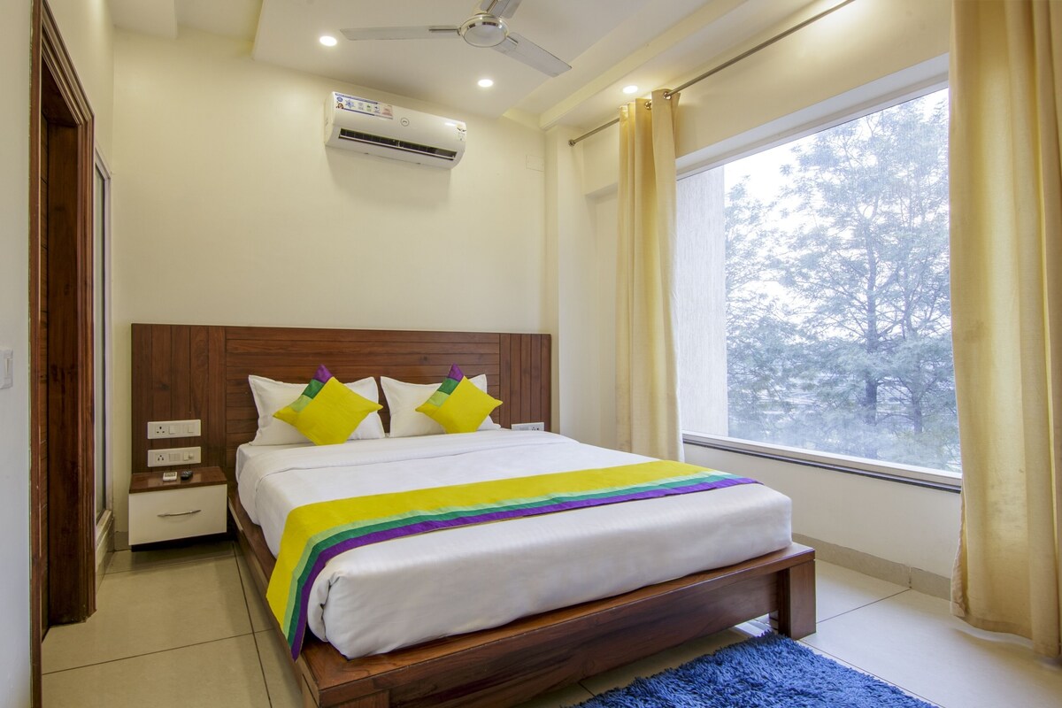 A bedroom with a wooden headboard, a bed with a rainbow-colored blanket, a ceiling fan, an air conditioning unit, a nightstand, and a window with curtains.