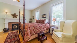 A four-poster bed with a red patterned bedspread, a small wooden stool, a cream-colored armchair, and a fireplace with a star decoration above it.